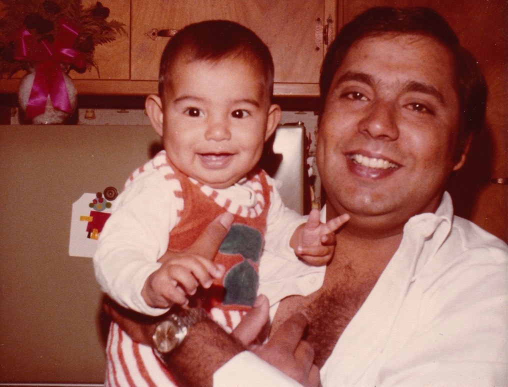 My father holding me as a baby, both of us laughing, in a kitchen — early 1980s.